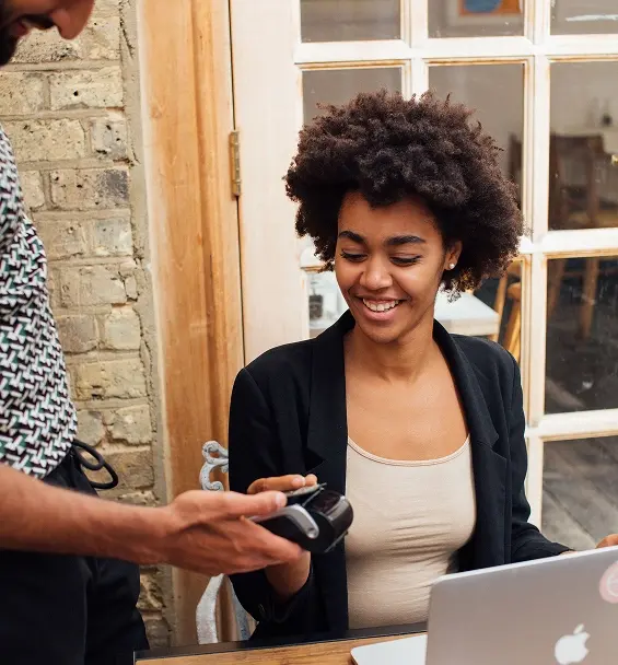 Smiling customer seated at a café table with a laptop, inserting her card into a handheld card reader held by a server.