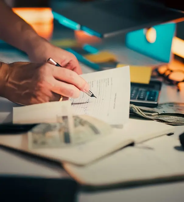 Close-up of a hand writing figures on a paper list beside a calculator, cash, sticky notes, and an open notebook on a desk.